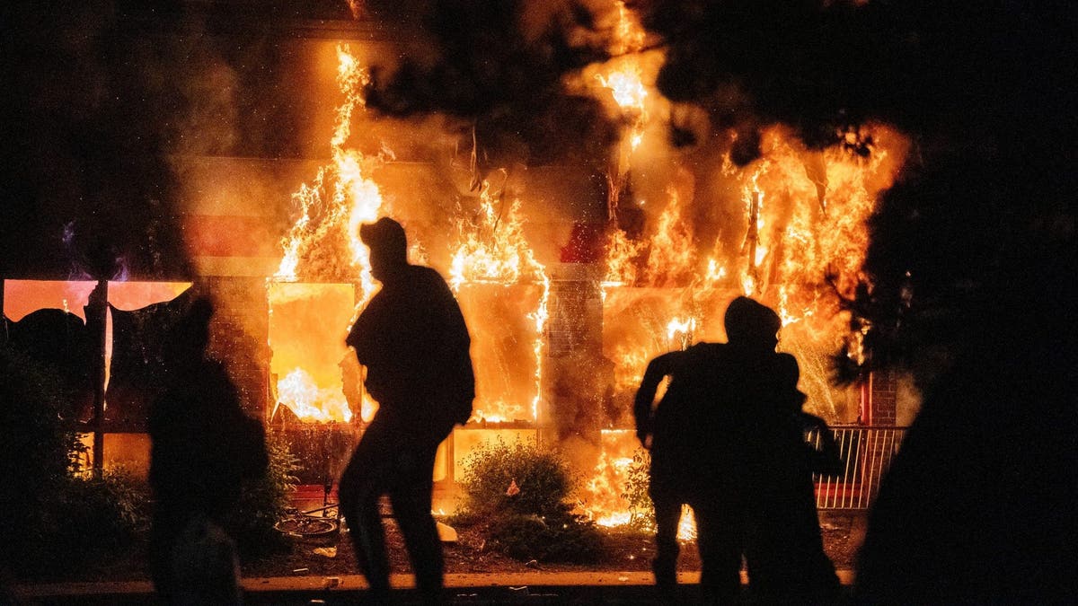 Flames engulf an AutoZone store as people move in front of the fire