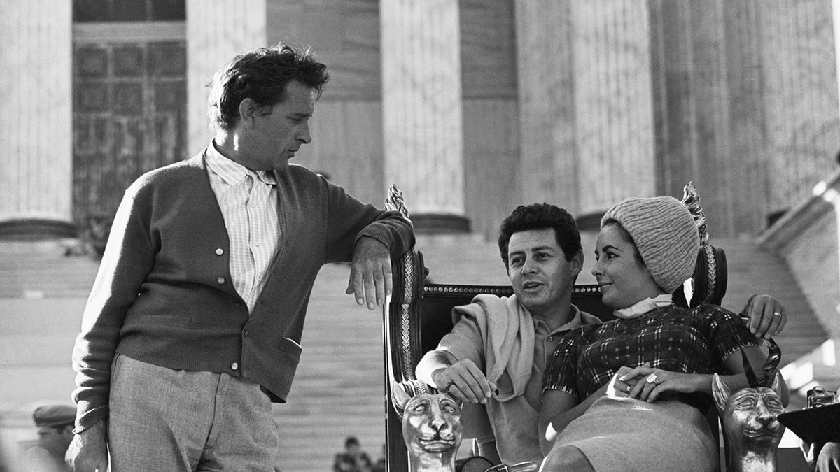 Richard Burton observing Elizabeth Taylor and Eddie Fisher as they sit together.